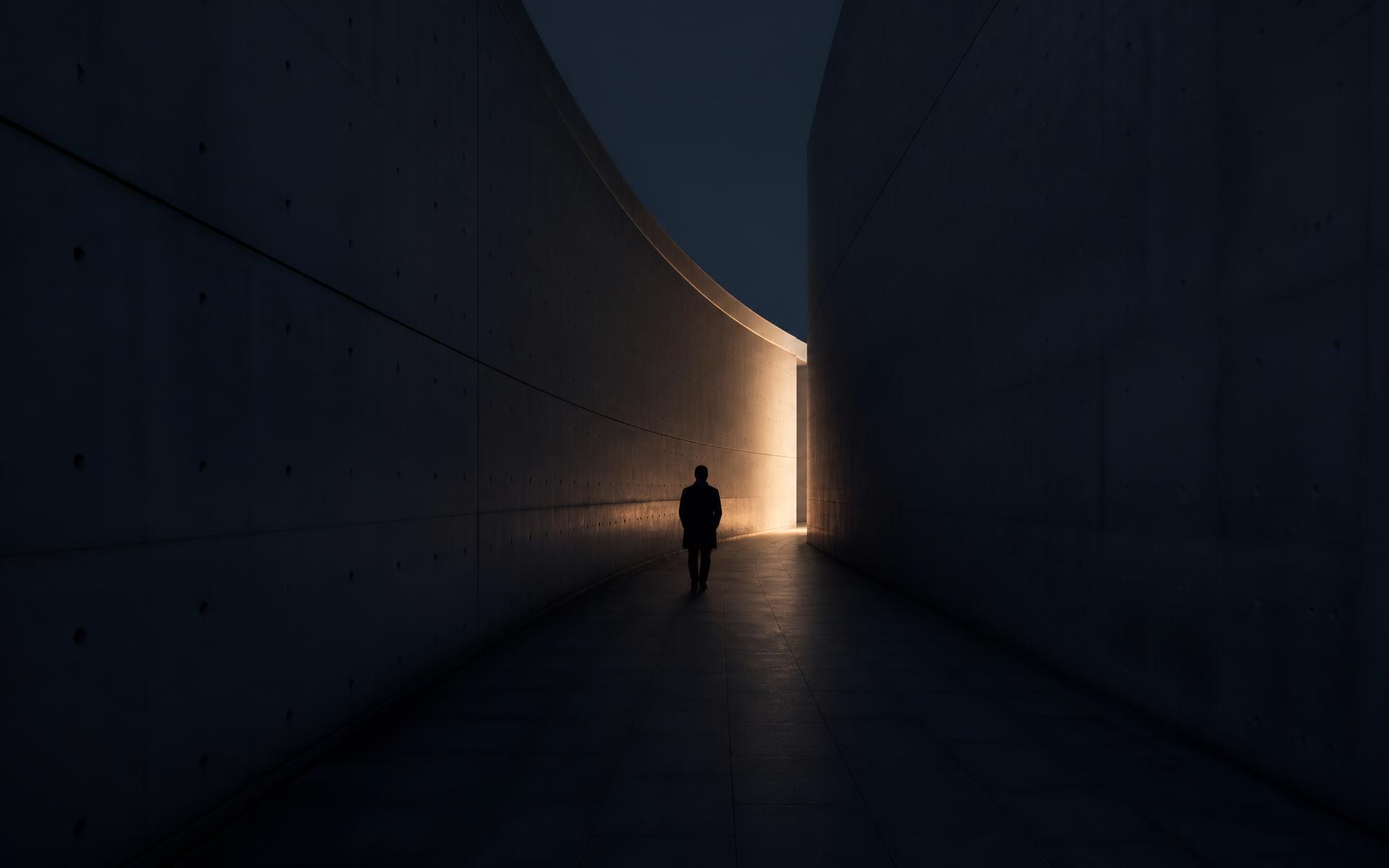 A single figure walking through a vast concrete corridor toward distant light